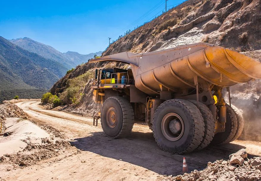 A large yellow dump truck drives on a dirt road next to a rocky hillside in a mountainous area, with blue sky and hills in the background. Dust rises from the ground beside the truck.