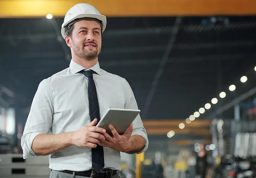 A man wearing a white safety helmet, dress shirt, and tie stands in an industrial setting, holding a tablet and looking ahead, with blurred machinery and lights in the background.