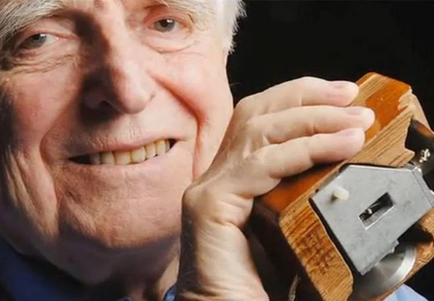 An elderly man smiles while holding a vintage wooden computer mouse close to his face, with a dark background.