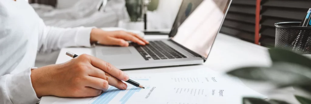 Una persona está sentada ante un escritorio con un ordenador portátil, mientras sostiene un bolígrafo y revisa un informe financiero impreso con tablas y gráficos. En el fondo se ven una planta y material de oficina.