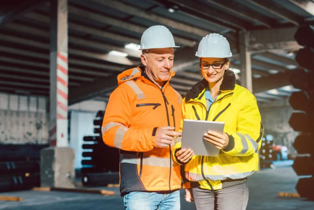 Two workers in safety helmets and high-visibility jackets stand in a warehouse, smiling while looking at a tablet. Sunlight streams in through windows, and stacks of materials are visible in the background.